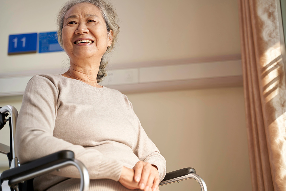An elderly woman with gray hair smiles while sitting in a wheelchair, illuminated by soft natural light in a bright room. There is a blue sign with the number "11" visible in the background.