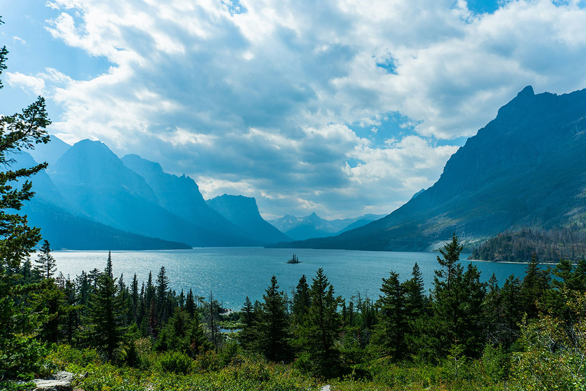 A scenic view of a lake surrounded by mountains and evergreen trees under a partly cloudy sky, with light reflecting off the water.