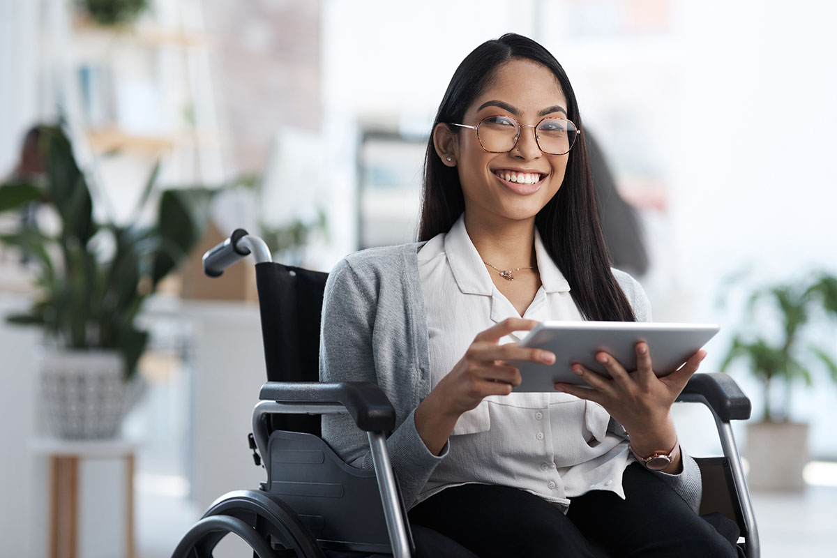 A smiling young woman in a wheelchair holds a tablet, dressed in a button-up shirt and a light cardigan, with indoor plants in the background.