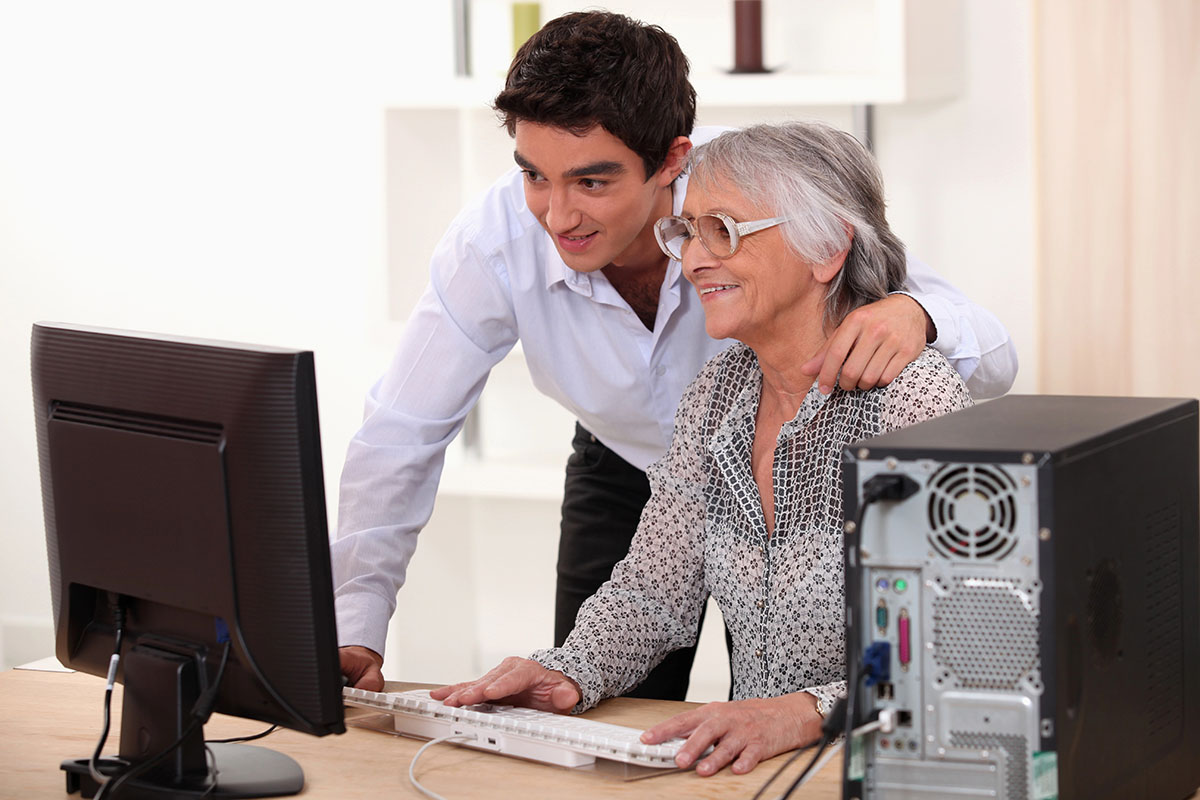 A young man leans over a keyboard to assist an older woman seated at a computer, both smiling as they engage with the screen in a bright, modern room.