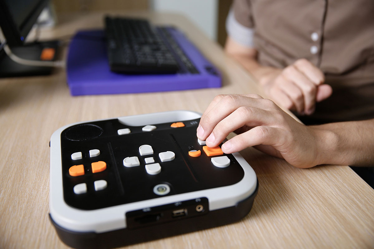 A person using a device with multiple buttons, including orange and white controls, on a wooden desk, with a computer keyboard in the background.