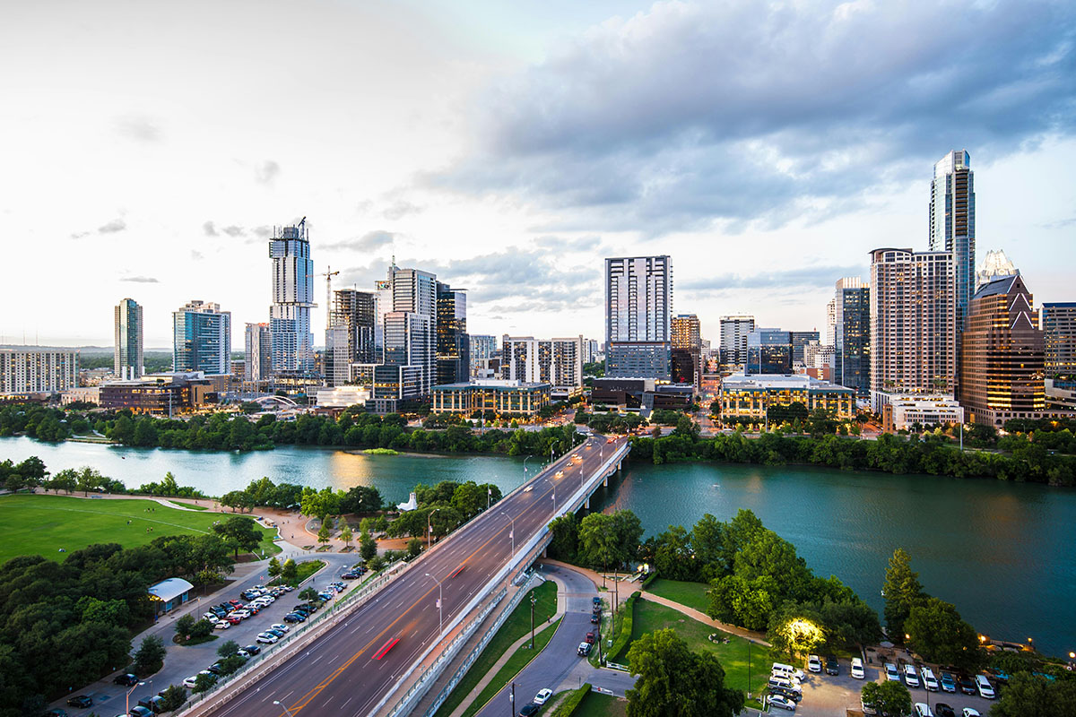 Aerial view of a city skyline with modern high-rise buildings reflecting in a river, surrounded by green parks and a busy roadway. The sky is partly cloudy, adding a dramatic backdrop.