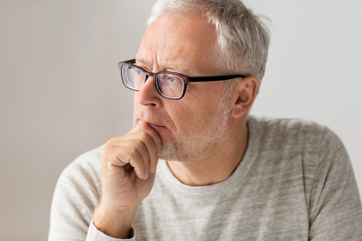 A thoughtful older man with gray hair and glasses rests his chin on his hand, looking off to the side against a soft, neutral background.