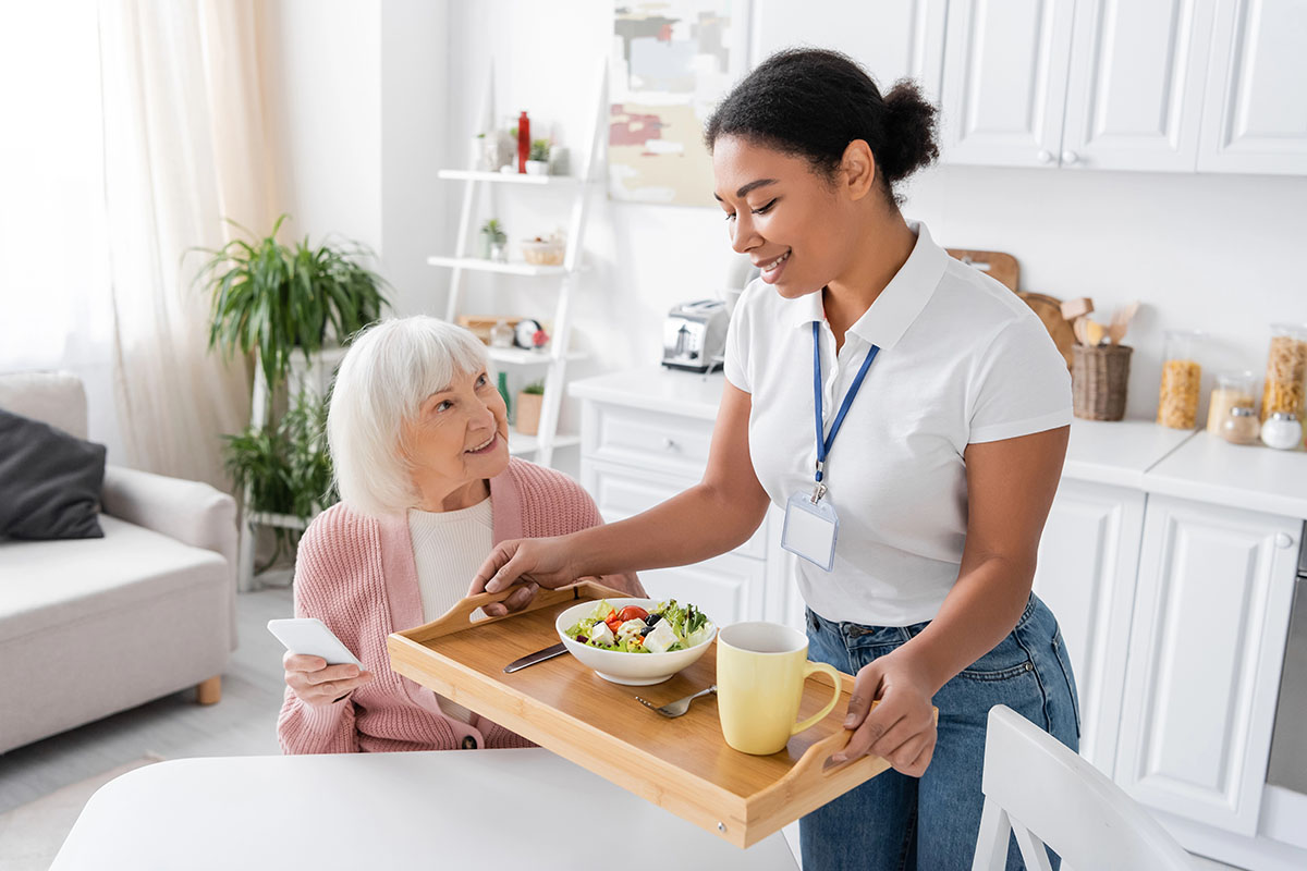 A caregiver serves a meal on a wooden tray to an elderly woman sitting at a table in a bright kitchen. The tray includes a bowl of salad and a yellow mug. The woman smiles at the caregiver, who is wearing a polo shirt and a name tag.