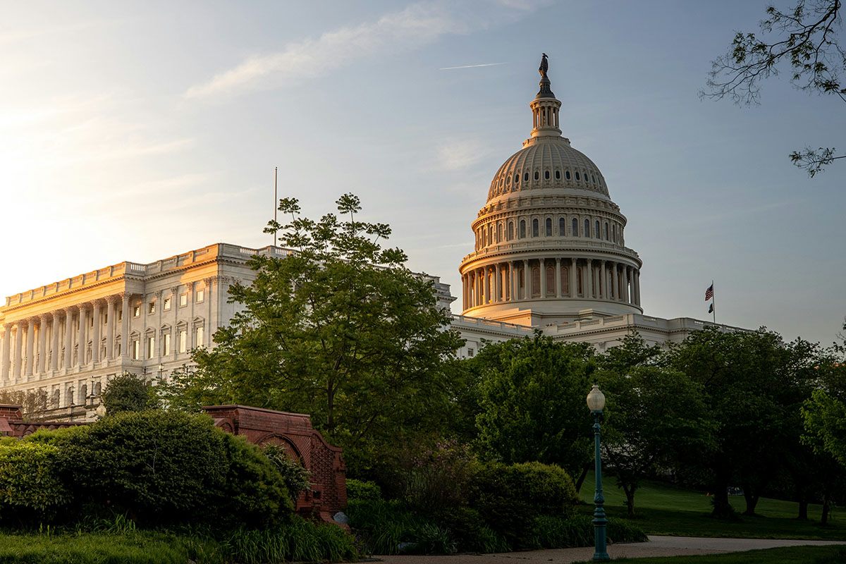 The U.S. Capitol building, with its iconic dome topped by a statue, is illuminated by a warm sunset glow, surrounded by lush trees and greenery.