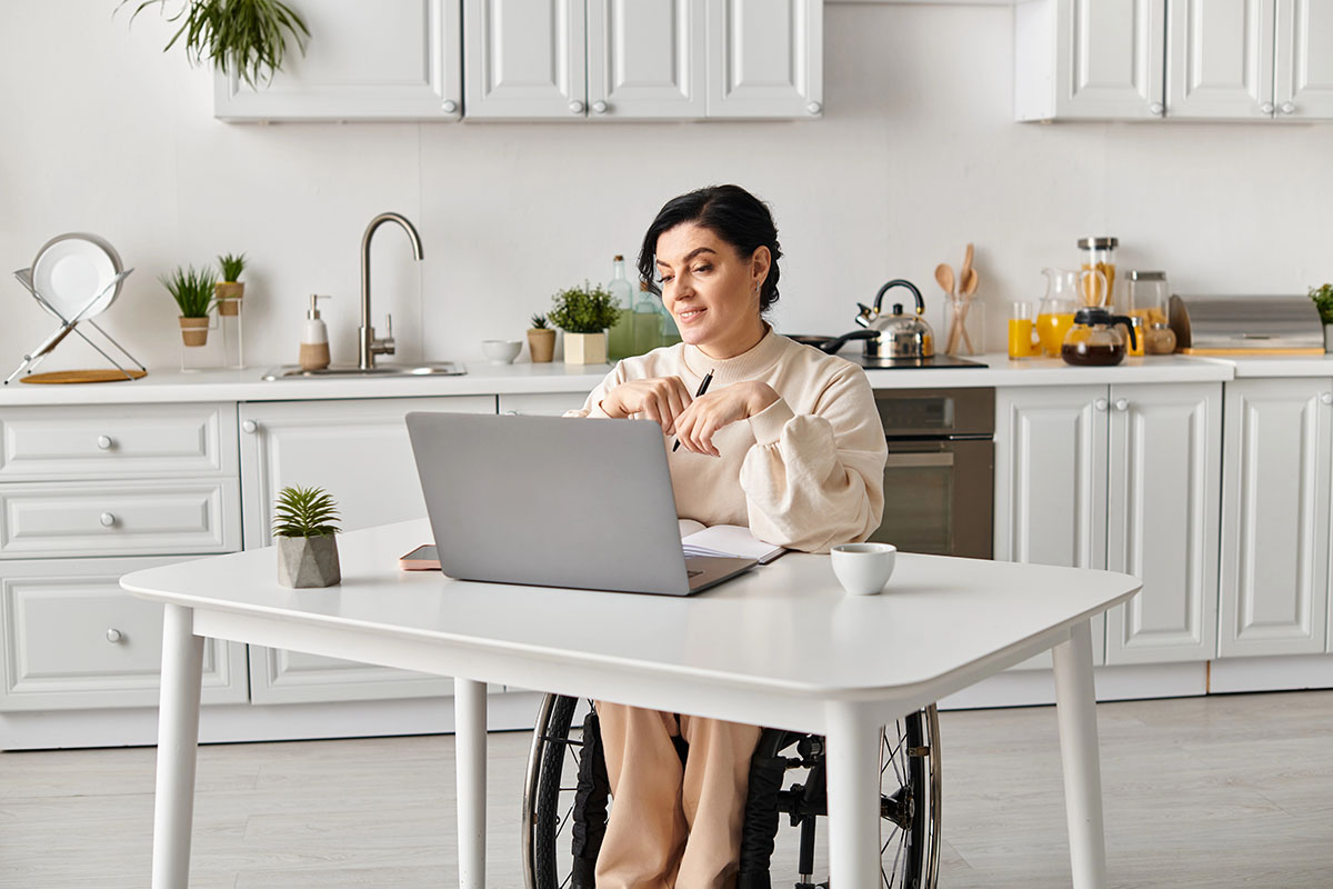 A woman in a wheelchair sits at a white table in a bright kitchen, looking at her laptop. She is wearing a cream-colored sweater and appears to be engaged in conversation while holding a pen. A small potted plant and a cup are on the table, with kitchen items in the background.