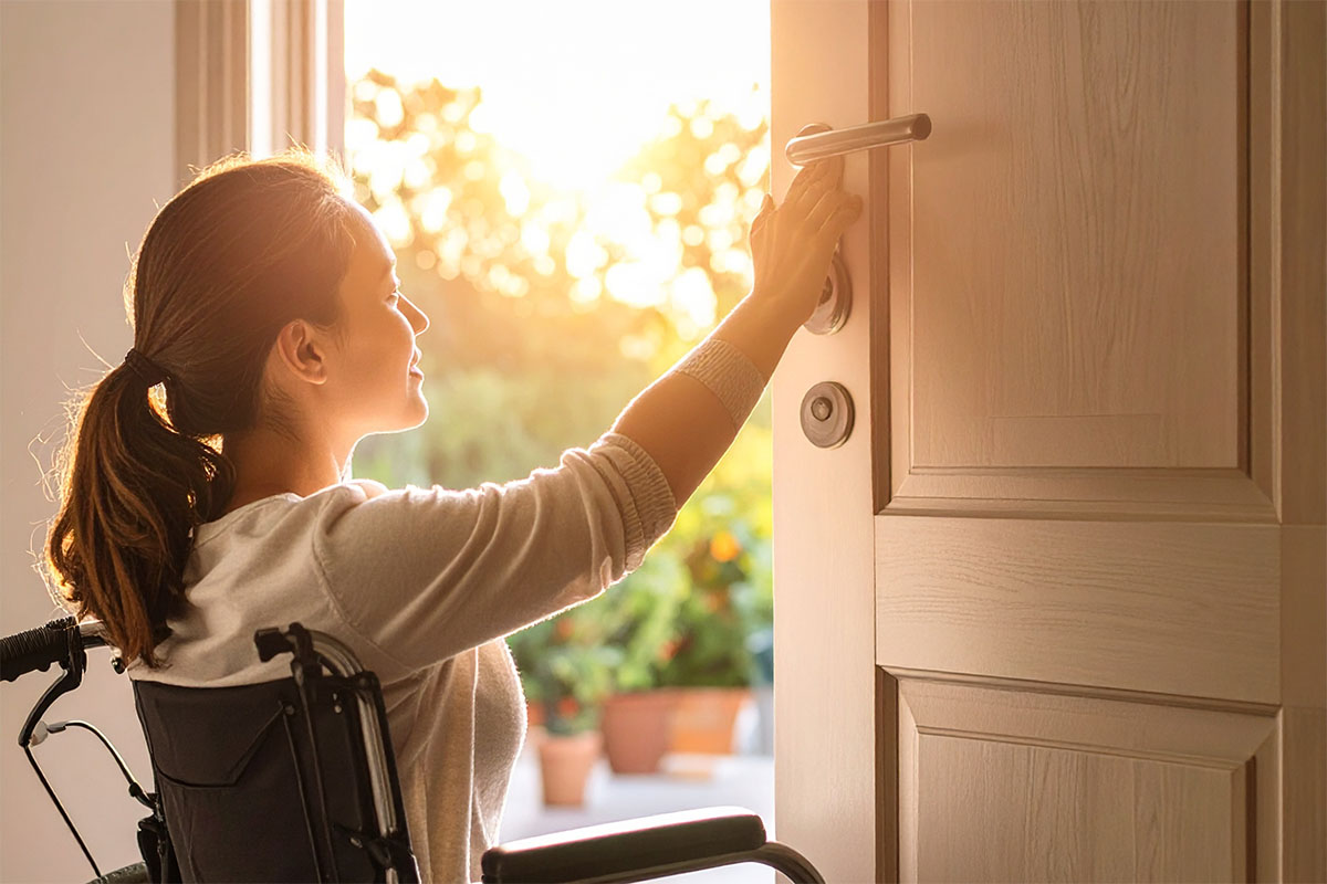 A woman in a wheelchair reaches for a doorknob, poised to open a door, with sunlight streaming in from behind her.