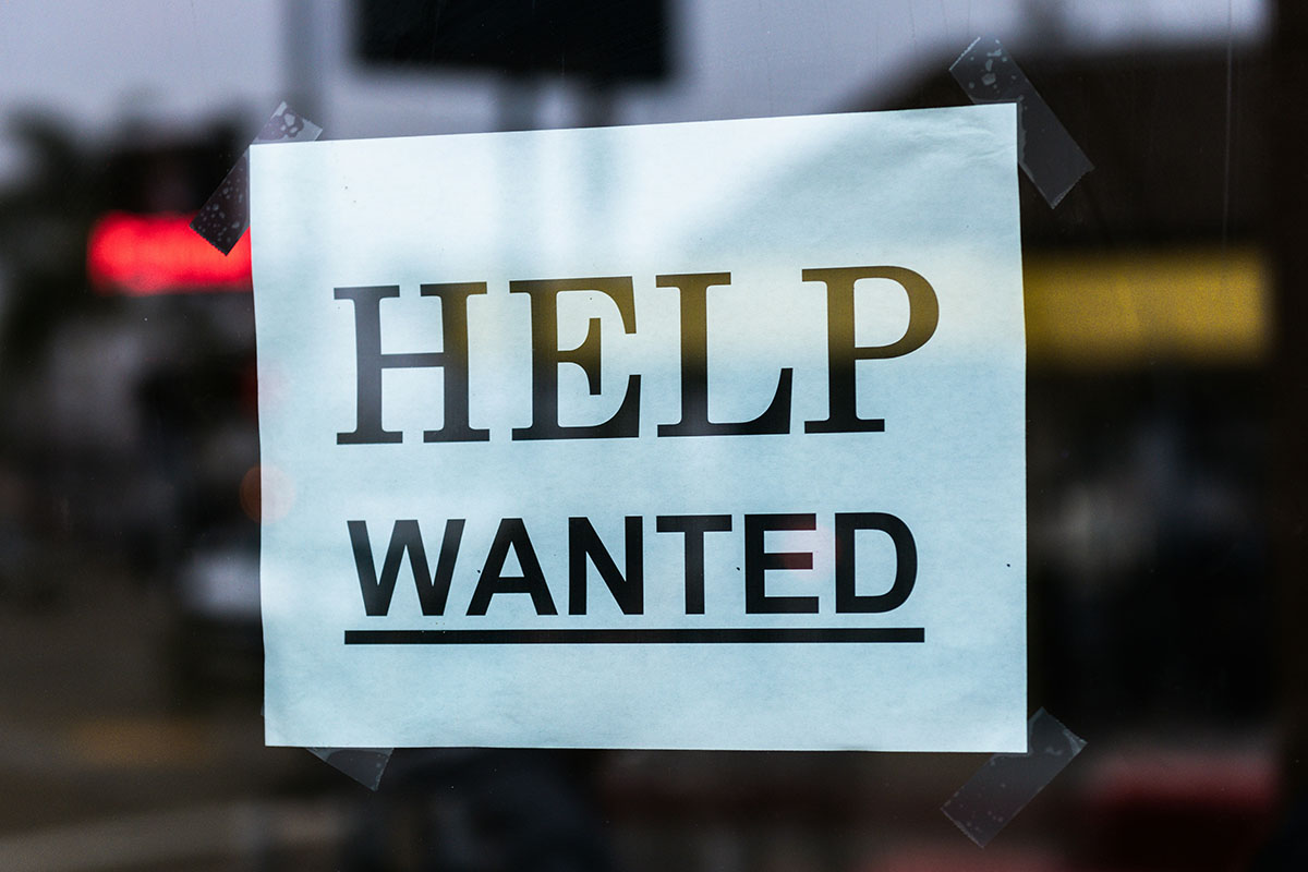 A blue sign taped to a window reads "HELP WANTED" in bold black letters.