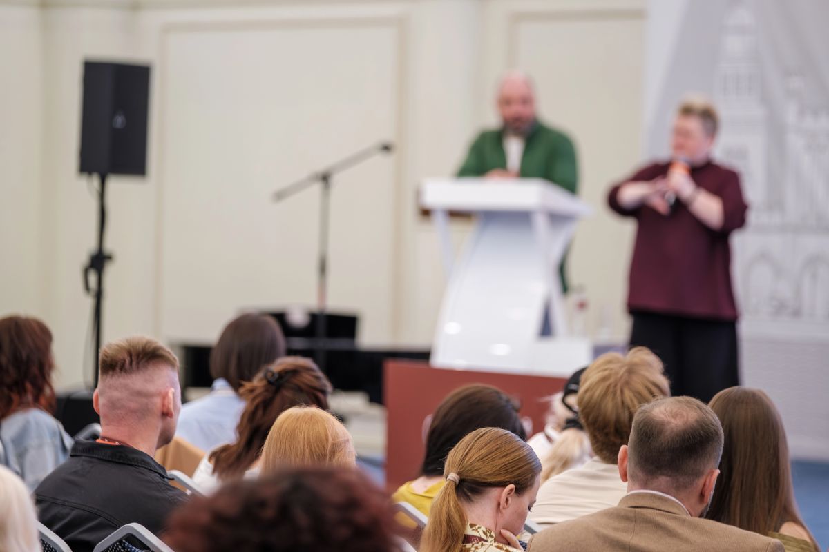 An audience listens to a speech with a speaker and sign language interpreter.