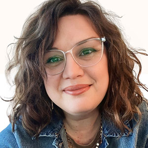 A woman with curly brown hair and green-tinted glasses smiles at the camera, wearing a denim jacket over a light top.