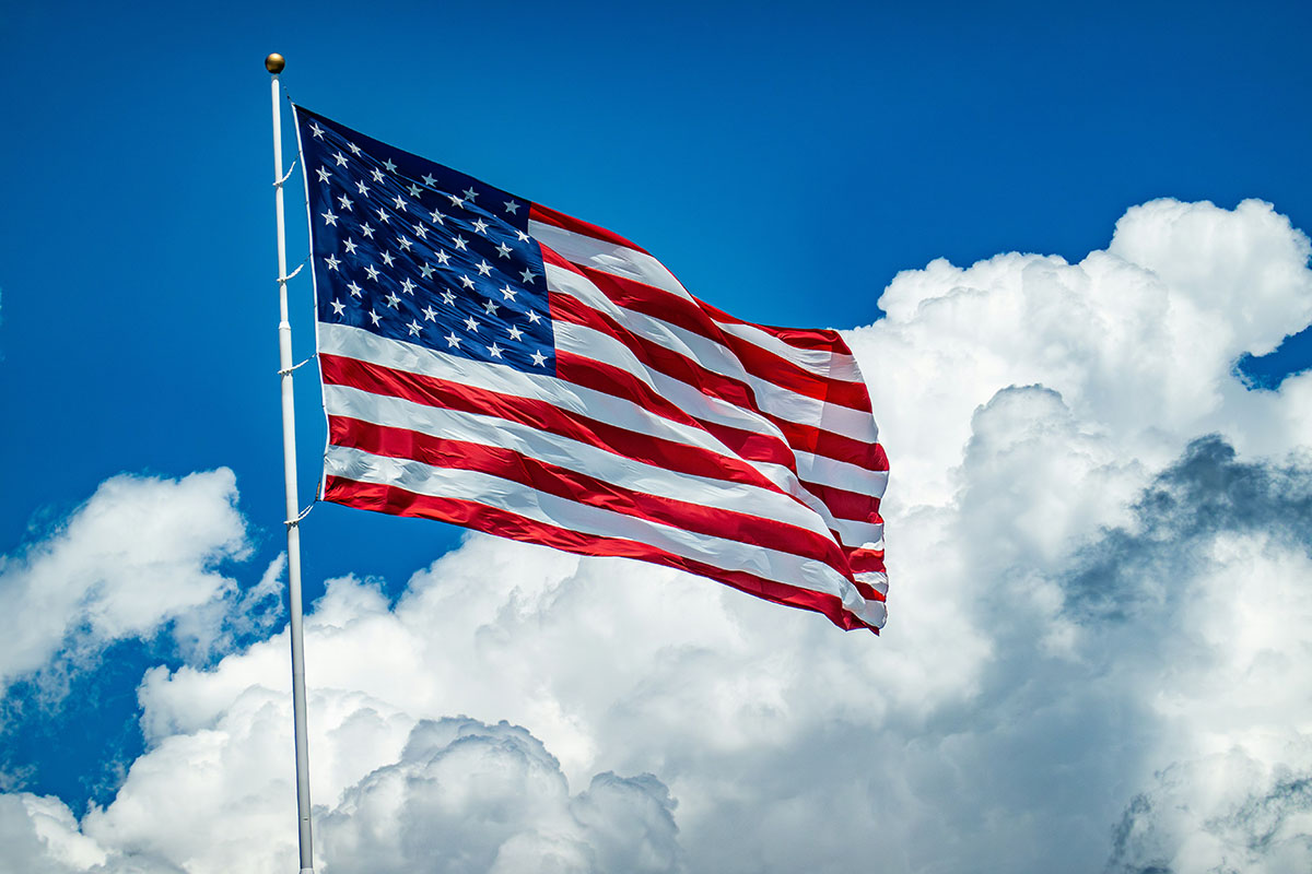 A U.S. flag with stars and stripes waving against a clear blue sky and fluffy white clouds.